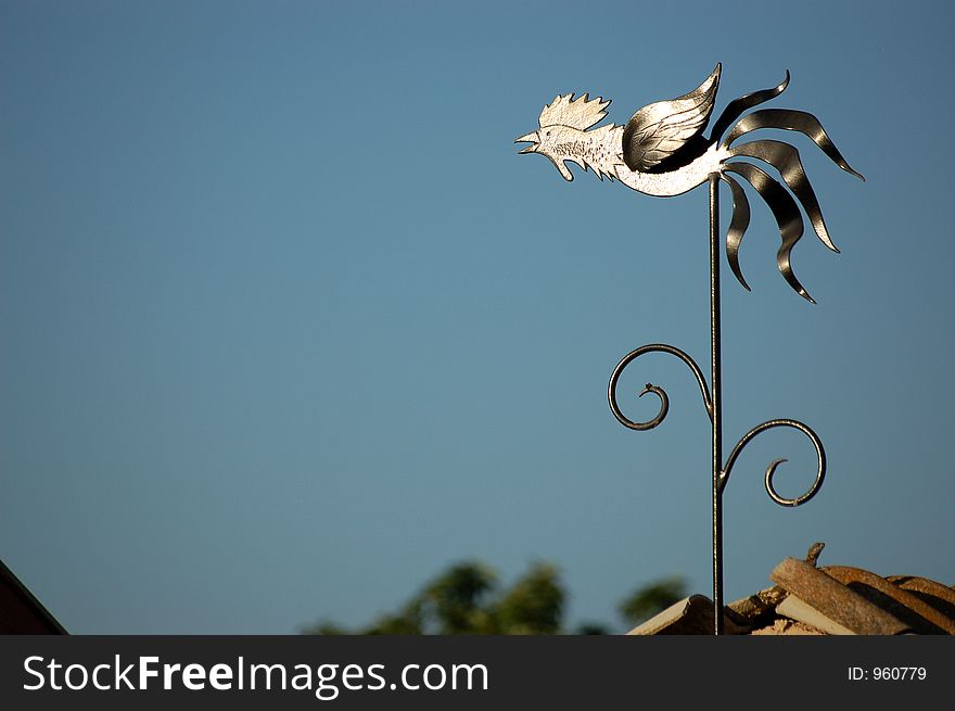 A metal chicken pictured on top of a roof, calling away, shining in the sun. A metal chicken pictured on top of a roof, calling away, shining in the sun.