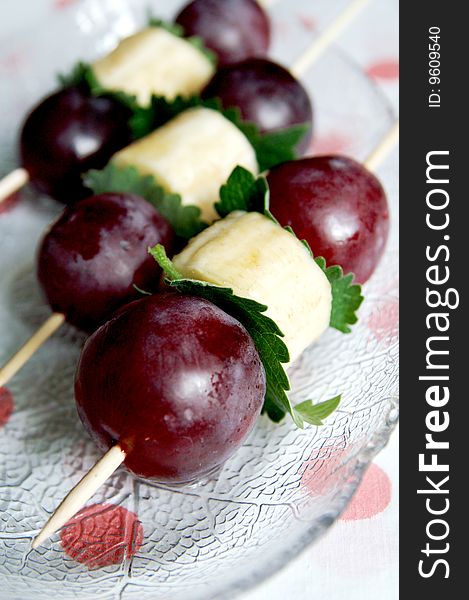 Red grapes and banana fruit kebab with mint leaves on wooden sticks, blur background, vertical, close-up