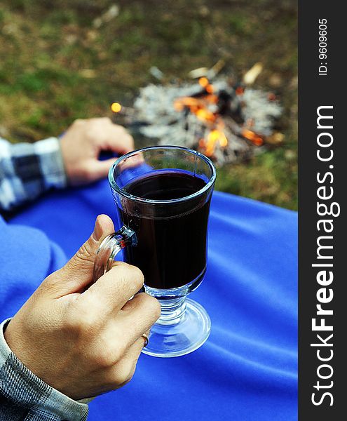 A mug with hot mulled wine in a hand of a man sitting near camp-fire under blue blanket at a picnic, blur background. A mug with hot mulled wine in a hand of a man sitting near camp-fire under blue blanket at a picnic, blur background