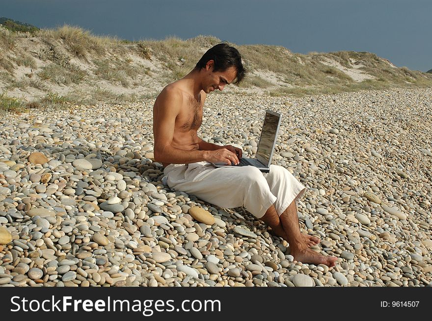 A young man sited on the beach working with a laptop. A young man sited on the beach working with a laptop