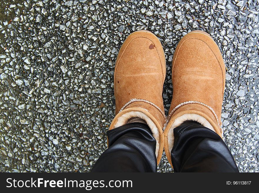 Feet in brown fleece lined boots standing on pavement from above. Feet in brown fleece lined boots standing on pavement from above.