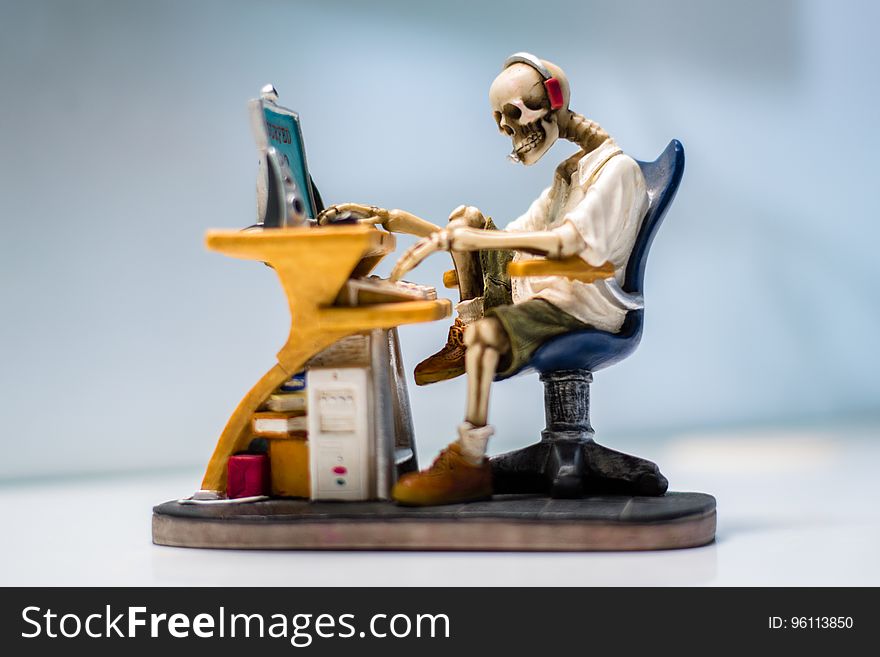 Skeleton (sculpture) wearing headphones sitting in a black swivel chair working the keyboard of a computer on a yellow desk, white and pale blue background. Concept of computers killing us. Skeleton (sculpture) wearing headphones sitting in a black swivel chair working the keyboard of a computer on a yellow desk, white and pale blue background. Concept of computers killing us.