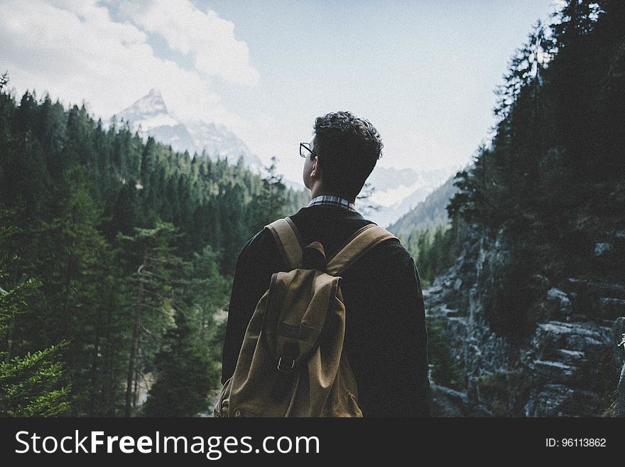 Back view of an anonymous adult male wearing a backpack, standing at a valley vista viewing the mountain landscapes that surround him. Back view of an anonymous adult male wearing a backpack, standing at a valley vista viewing the mountain landscapes that surround him.