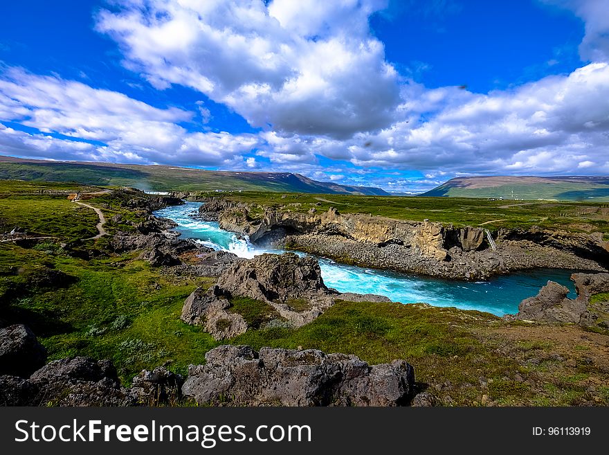 Aerial view of river with rocky banks through green field in countryside with blue skies and white clouds. Aerial view of river with rocky banks through green field in countryside with blue skies and white clouds.