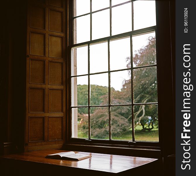 Open book on wooden table next to sunny window inside home. Open book on wooden table next to sunny window inside home.