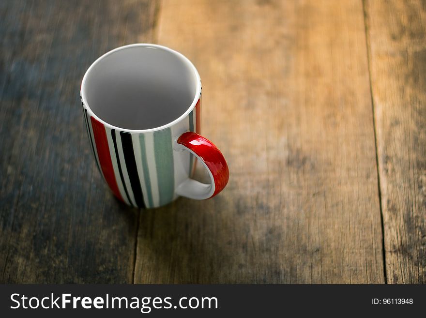 Empty mug with green, black, white and red vertical stripes, on a table made of wooden planking partly (sanded) bleached. Empty mug with green, black, white and red vertical stripes, on a table made of wooden planking partly (sanded) bleached.