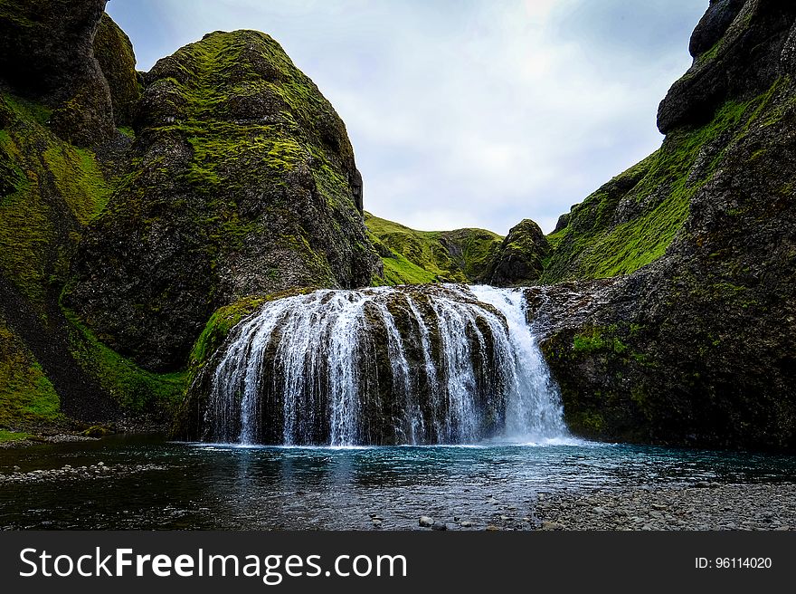 Waterfalls over green cliffs into lake or stream with cloudy skies on sunny day.