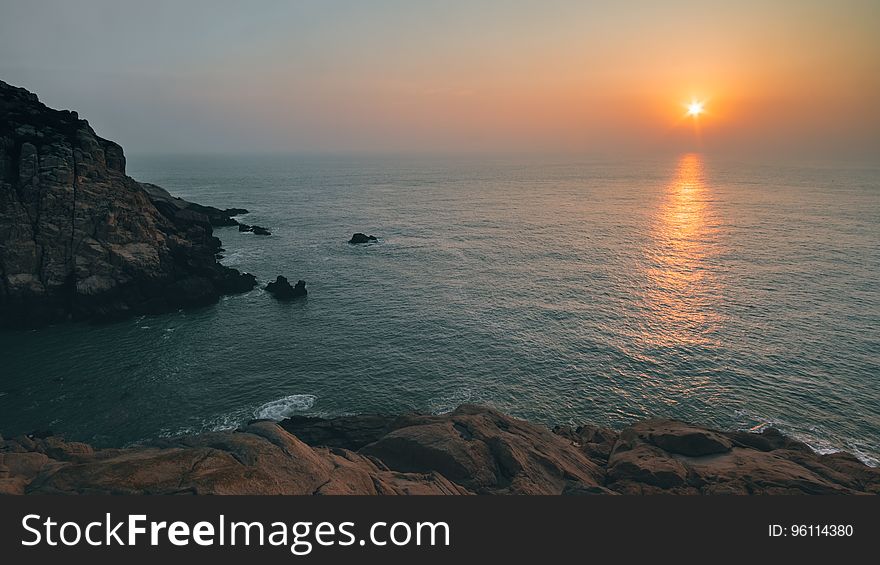 A view of a rocky coast with the sun rising in the distance. A view of a rocky coast with the sun rising in the distance.