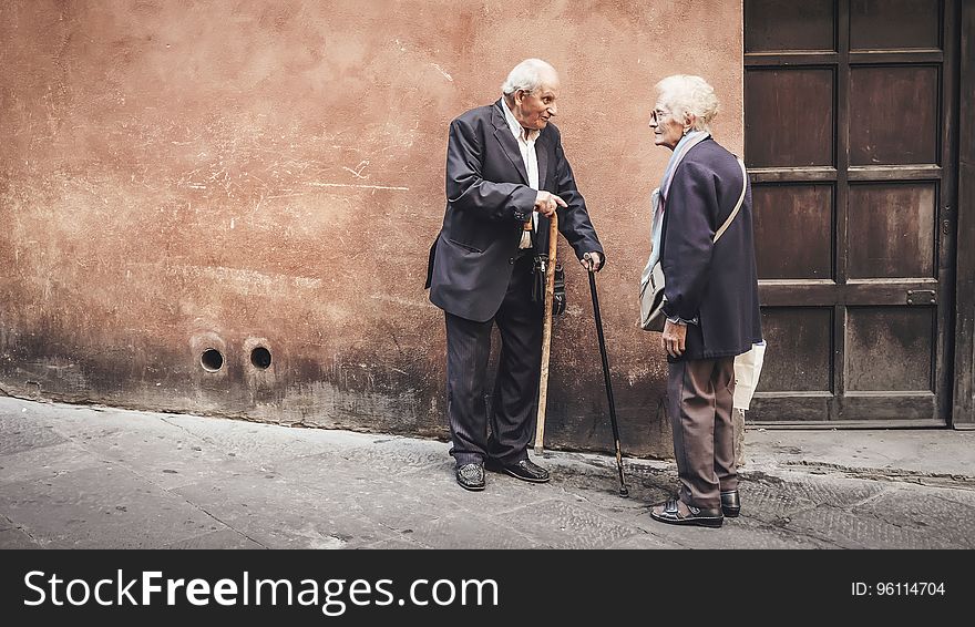 Elder man and woman with cane standing outside building having conversation on sunny day. Elder man and woman with cane standing outside building having conversation on sunny day.