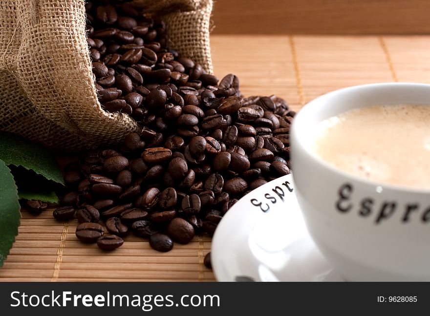 A bunch of coffee beans, falling out of a sack and a cup of fresh coffee on a wooden background