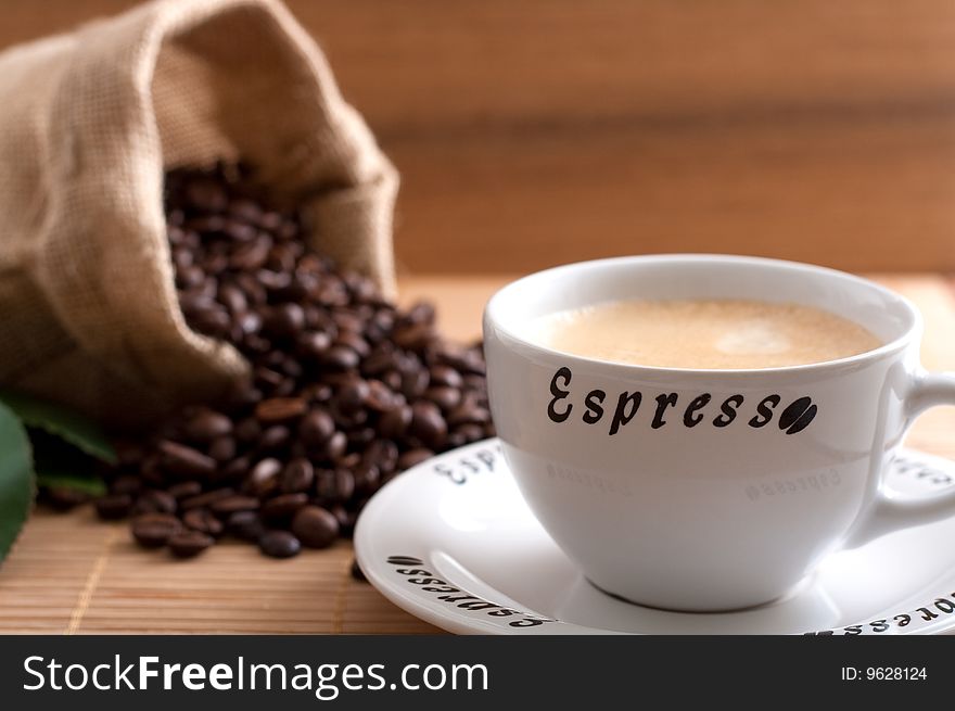 A bunch of coffee beans, falling out of a sack and a cup of fresh coffee on a wooden background