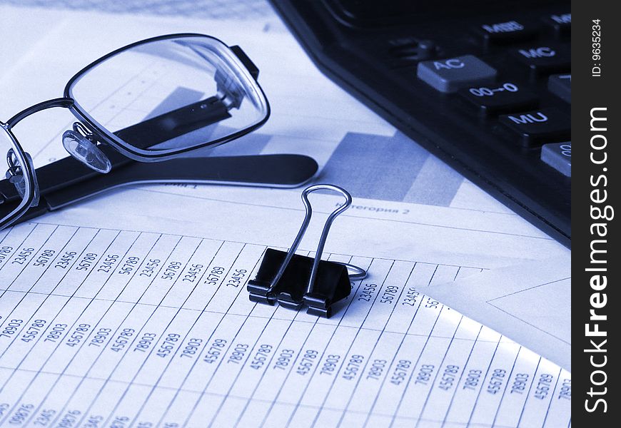 Glasses, calculator and binder clip on financial documents. Toned blue. Shallow DOF.