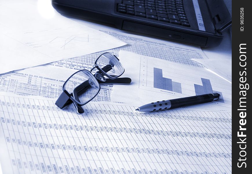 Laptop, glasses and pen on financial documents. Toned blue. Shallow DOF.