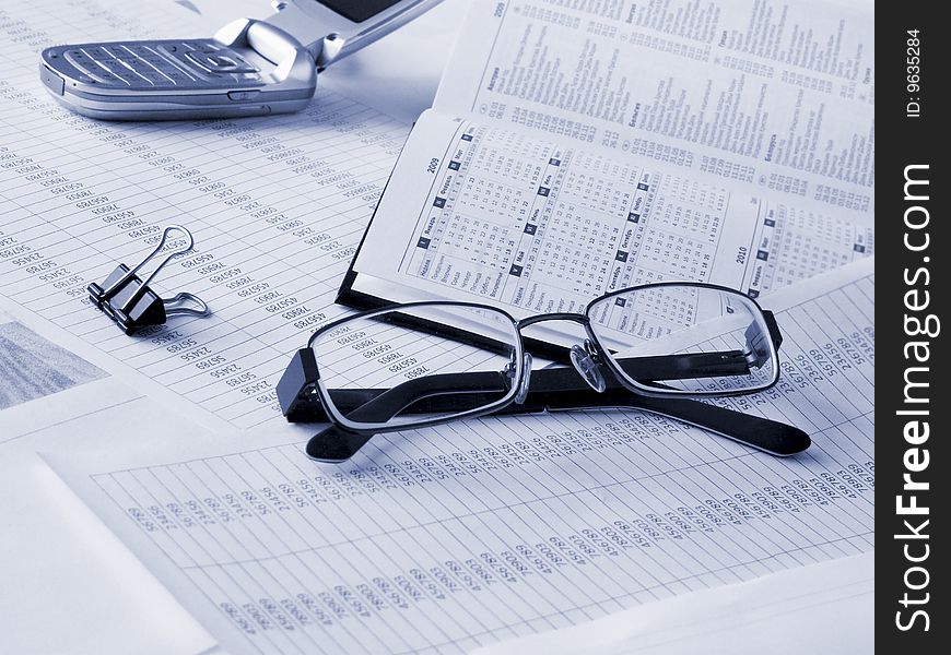 Glasses, daily book and mobile phone on financial documents. Toned blue. Shallow DOF.