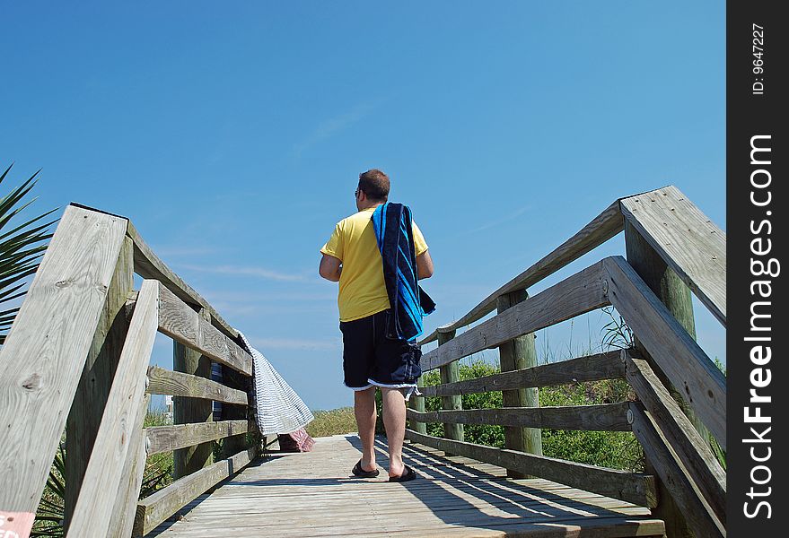 A vacationing man, carrying his towel over his shoulder, is walking along a boardwalk on his way to the beach. A vacationing man, carrying his towel over his shoulder, is walking along a boardwalk on his way to the beach.