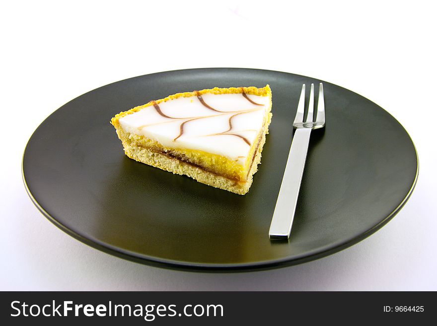 Delicious looking iced bakewell tart on a black plate with a fork and a plain background