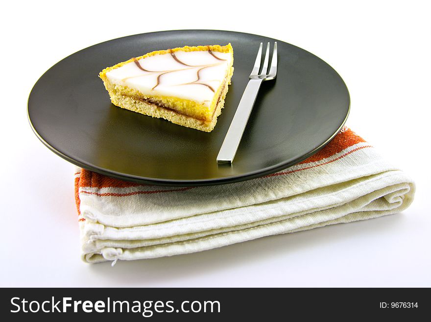 Delicious looking iced bakewell tart on a black plate with a fork and a plain background