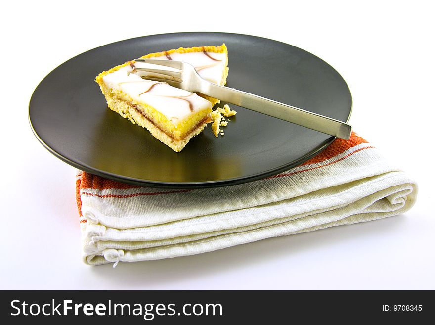 Delicious looking iced bakewell tart on a black plate with a fork and a plain background