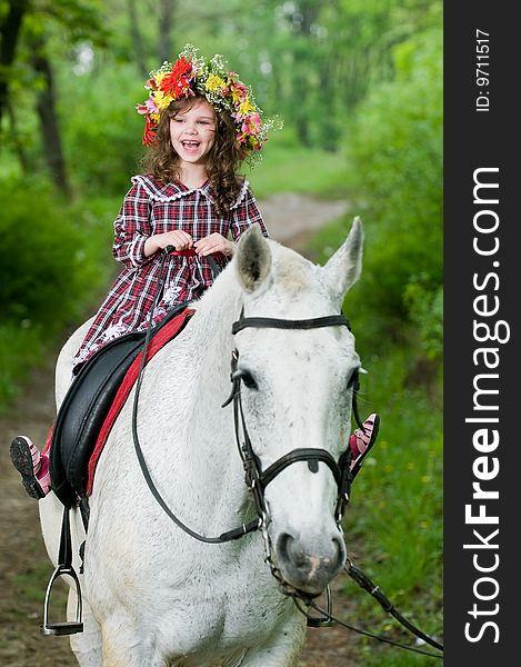 Laughing little girl in floral wreath riding horse in the forest