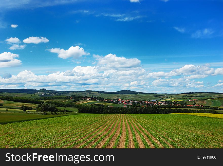 Rural landscape with a cozy village among the hills, fields and meadows under cloudy blue sky. Rural landscape with a cozy village among the hills, fields and meadows under cloudy blue sky