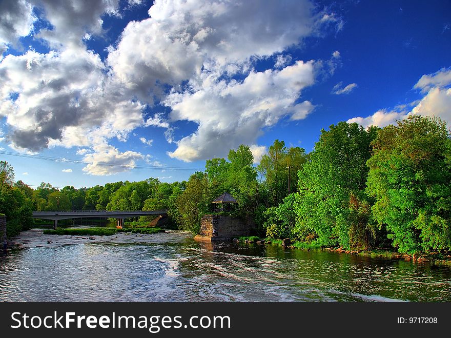 A river flows towards a bridge under puffy clouds on a warm afternoon. A man can be seen in the distance fishing from the right bank. A river flows towards a bridge under puffy clouds on a warm afternoon. A man can be seen in the distance fishing from the right bank.