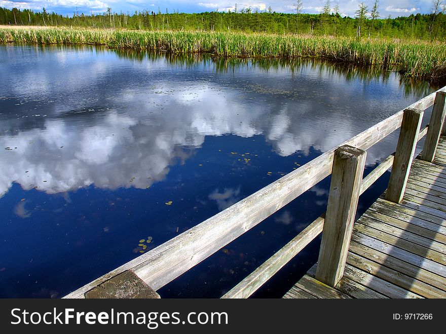 Afternoon clouds are reflected in a calm pond by a wooden boardwalk. Afternoon clouds are reflected in a calm pond by a wooden boardwalk.