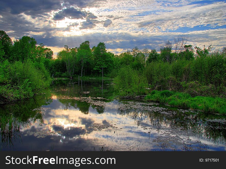 Clouds are reflected on the surface of a still pond as the sun is starting to shine through. Clouds are reflected on the surface of a still pond as the sun is starting to shine through.