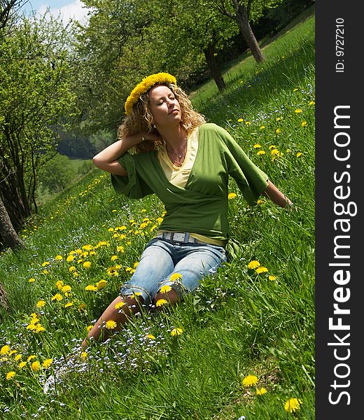 Curly girl with dandelion chain on head sitting on meadow in blossom