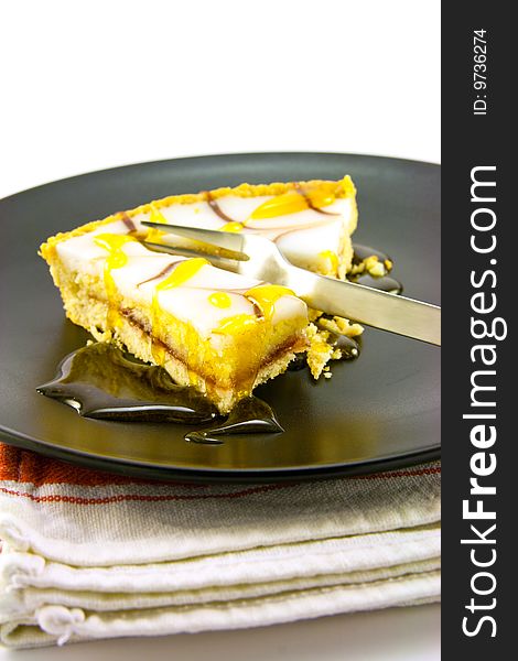 Delicious looking iced bakewell tart on a black plate with a treacle drizzle and a fork on a plain background