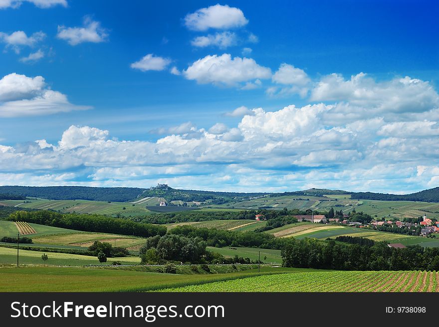 Rural landscape with a cozy village among the hills, fields and meadows under cloudy blue sky. Rural landscape with a cozy village among the hills, fields and meadows under cloudy blue sky