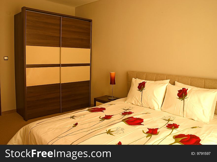 Warm bedroom with wardrobe and large bed with pillows and roses sheets. Warm bedroom with wardrobe and large bed with pillows and roses sheets.