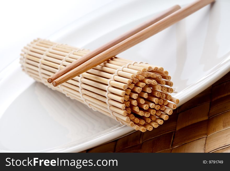 A tilted horizontal macro of a pair of chopsticks and a sushi rolling mat on a white plate. A tilted horizontal macro of a pair of chopsticks and a sushi rolling mat on a white plate