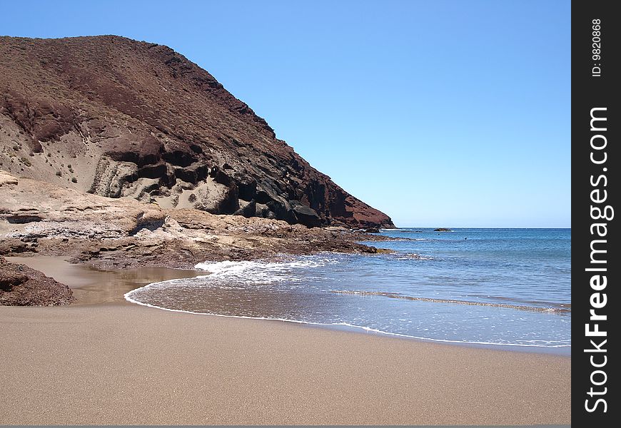 Waves gently lap the sandy shores of a quiet beach under a red cliff. Waves gently lap the sandy shores of a quiet beach under a red cliff