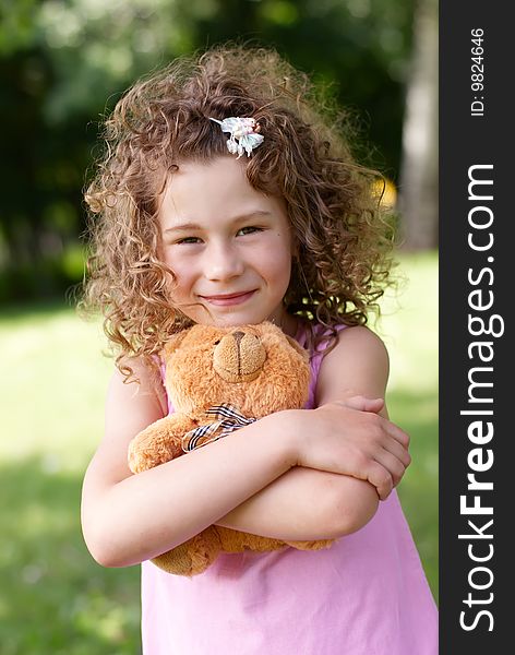 The beautiful girl of 7 years embraces a teddy bear on a green vegetative background in the summer. The beautiful girl of 7 years embraces a teddy bear on a green vegetative background in the summer