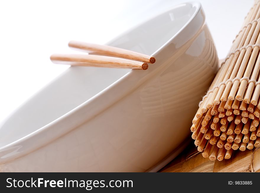 A tilted horizontal image of a pair of chopsticks on a white bowl with a sushi rolling mat on a weaved place mat