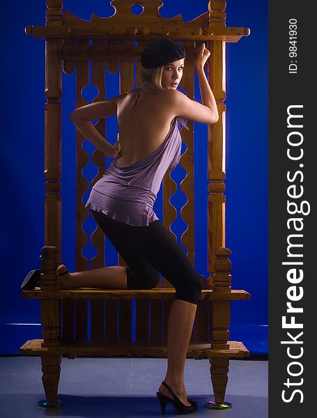 Beautiful young girl standing beside a chair in purple T-shirt, black cap and the bridge on a blue background. Beautiful young girl standing beside a chair in purple T-shirt, black cap and the bridge on a blue background