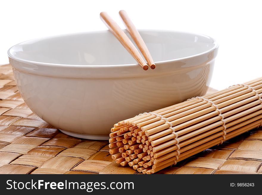 A horizontal image of a pair of chopsticks on a white bowl with a sushi rolling mat on a weaved place mat