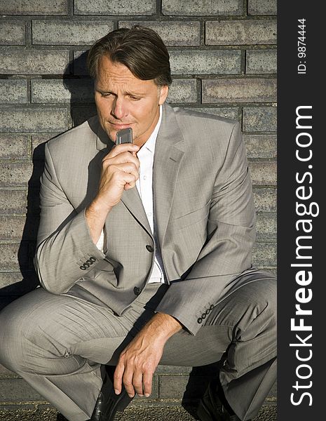 A depressed and lonely looking forties businessman is crouching down against a brick wall and holding his cellphone after hearing bad news. A depressed and lonely looking forties businessman is crouching down against a brick wall and holding his cellphone after hearing bad news.