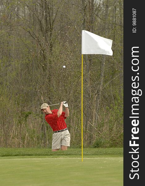 A man blasts out of a bunker kicking up sand. A man blasts out of a bunker kicking up sand