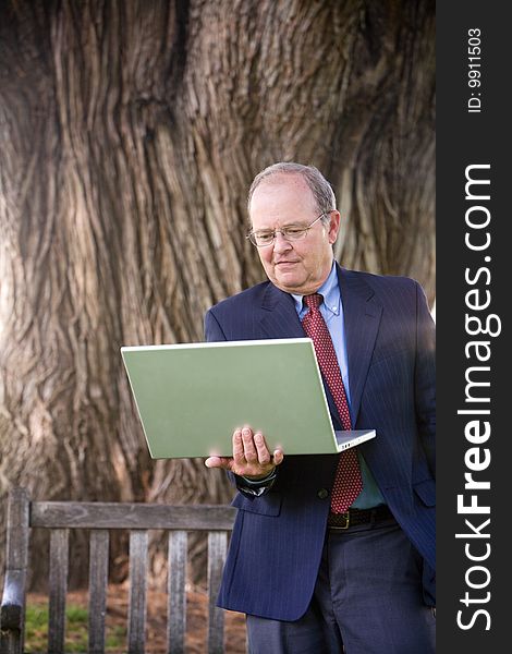 An older business man stands in the park while working on his laptop computer. An older business man stands in the park while working on his laptop computer