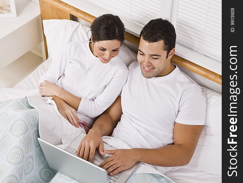 An attractive young couple lying in bed together and viewing a laptop screen.  They are both smiling. Horizontally framed shot. An attractive young couple lying in bed together and viewing a laptop screen.  They are both smiling. Horizontally framed shot.