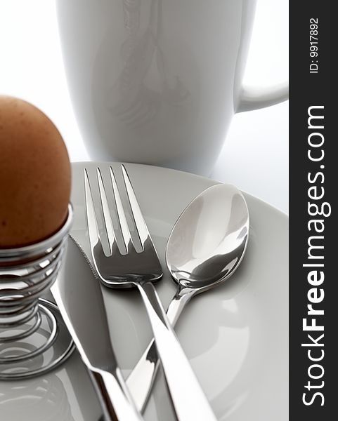 Egg, knife, fork, spoon on a white plate against a white cup. Egg, knife, fork, spoon on a white plate against a white cup