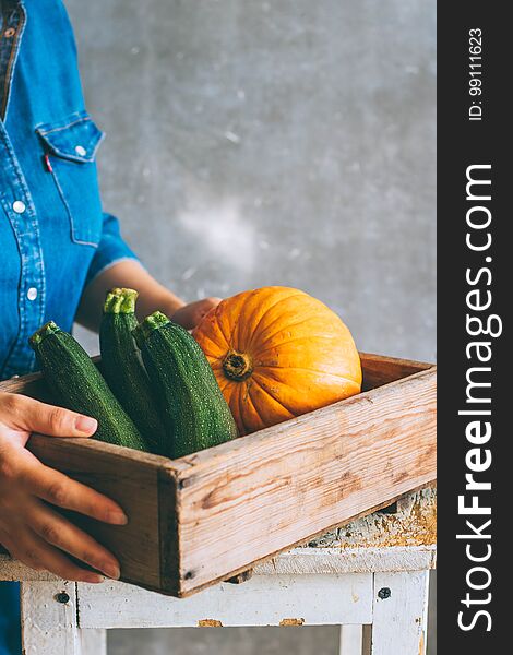 A girl in a blue dress is holding an autumn harvest in a wooden box, a pumpkin, a green zucchini on a gray background