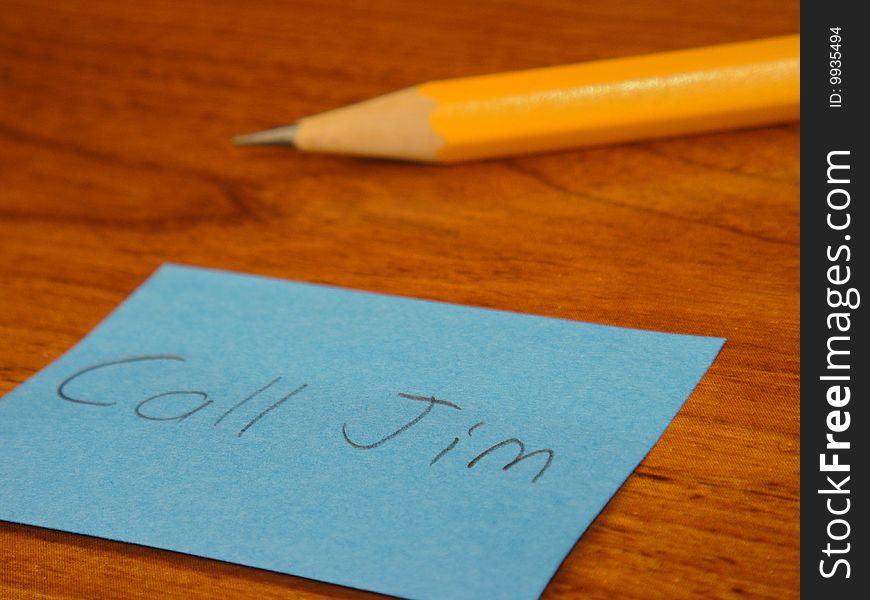 A sticky note that has Call Jim written on it with a pencil in the background on a desk. A sticky note that has Call Jim written on it with a pencil in the background on a desk.