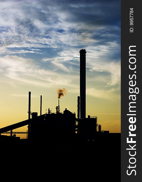 Black silhouette of an industrial facility against blue sky with some clouds backlit by sunset. Black silhouette of an industrial facility against blue sky with some clouds backlit by sunset