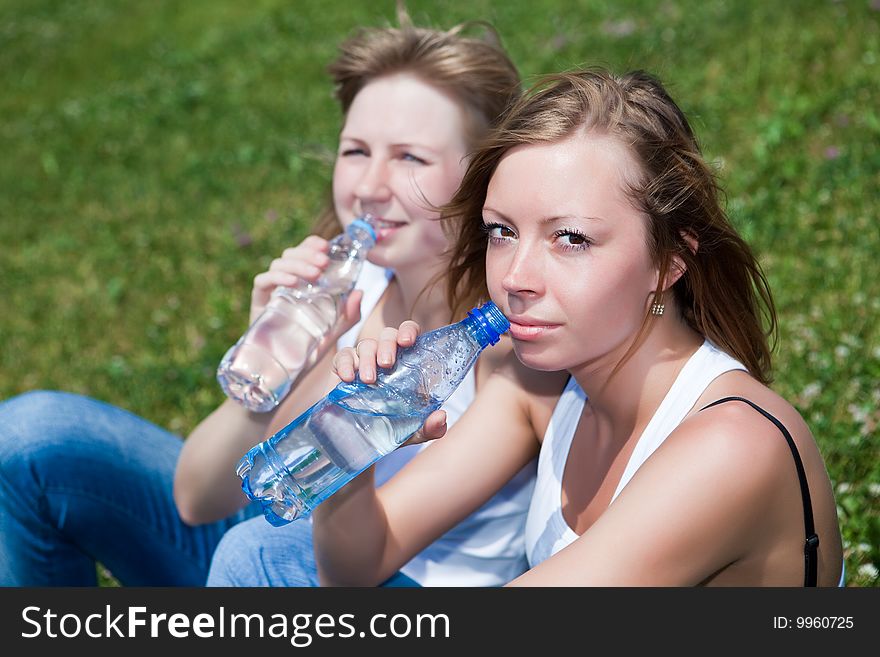 Two beautiful girls drink water sitting on a green grass. Two beautiful girls drink water sitting on a green grass