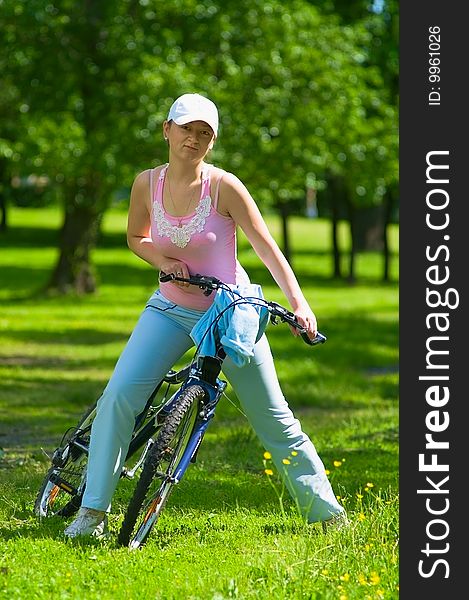 Young woman biker on the bicycle stand in the park. She going to ride on bicycle. It`s a hot summer. Young woman biker on the bicycle stand in the park. She going to ride on bicycle. It`s a hot summer.