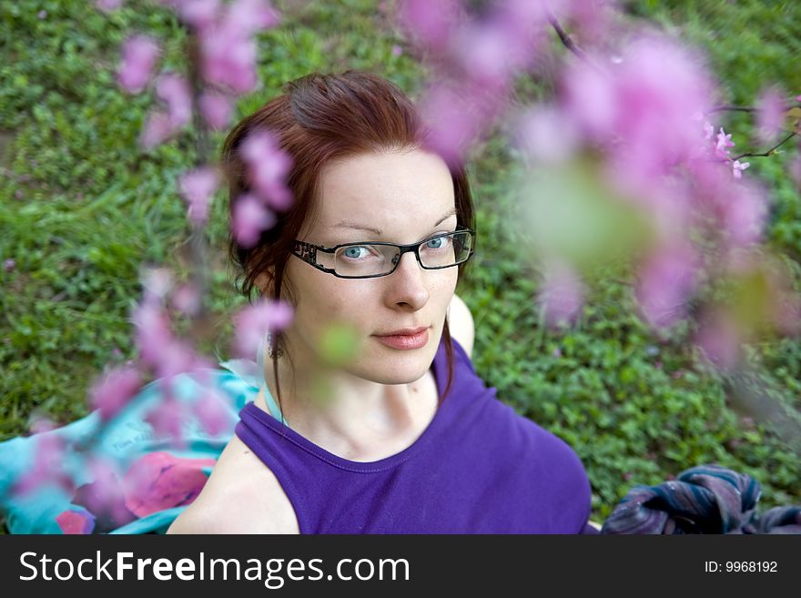 Young woman with glasses in park sitting on grass. Blured flowers in front. Young woman with glasses in park sitting on grass. Blured flowers in front.