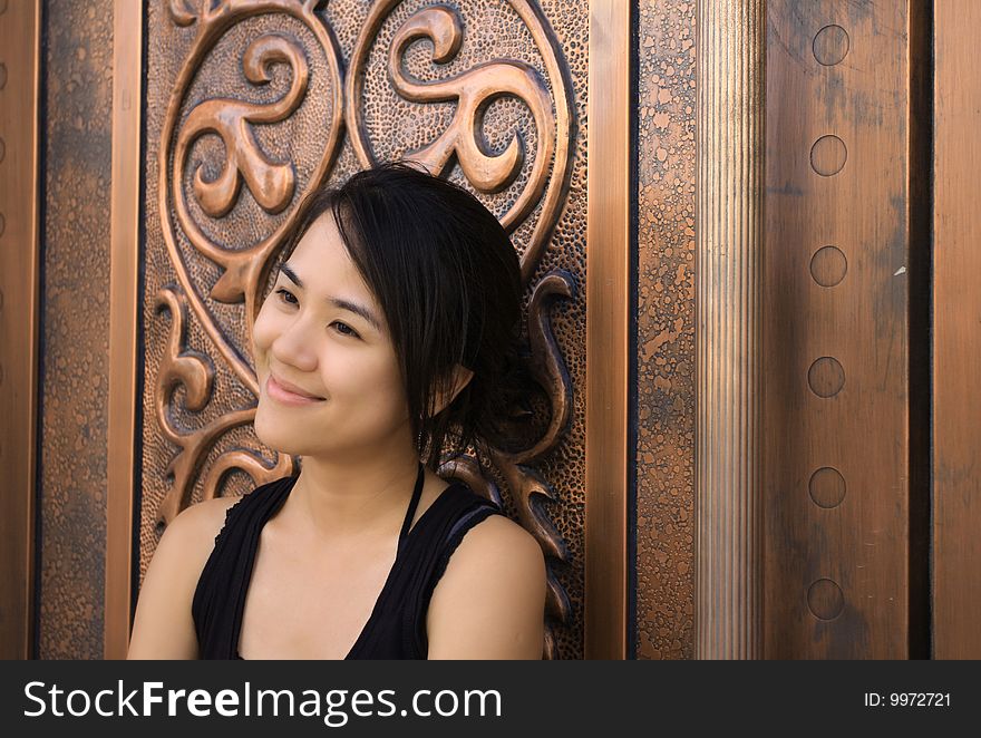 A young Chinese woman leaning on an old gloden cuprum door with a sweet smile.