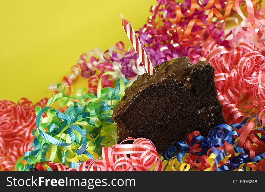 A piece of two layer chocolate cake with one candle with a party background of mutlicolored curly ribbons, horizontal with copy space. A piece of two layer chocolate cake with one candle with a party background of mutlicolored curly ribbons, horizontal with copy space